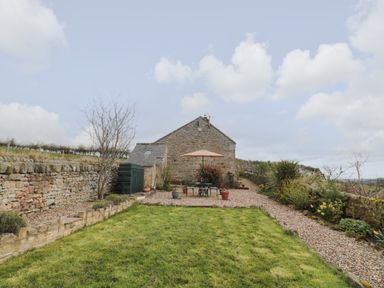 An outdoor area with a stone wall a grass lawn a gravel pathway and a stone building with a patio table and umbrella at Healey Farm Cottage in Rothbury