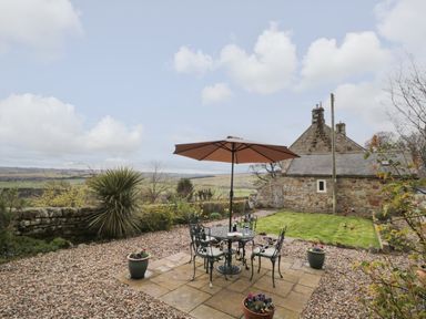 A garden patio with a table and four chairs under an umbrella and a stone building in the background at Healey Farm Cottage in Rothbury