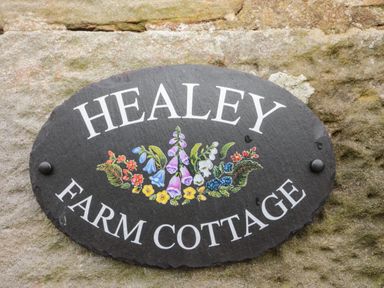 A black oval sign with white text Healey Farm Cottage and painted flowers on a stone wall at Healey Farm Cottage in Rothbury