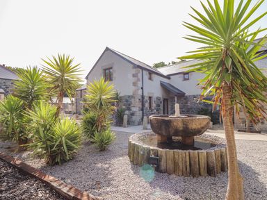 An outdoor garden with palm-like plants gravel ground and a stone water fountain in front of a stone and white plaster house at Polurrian House in Cury near Mullion