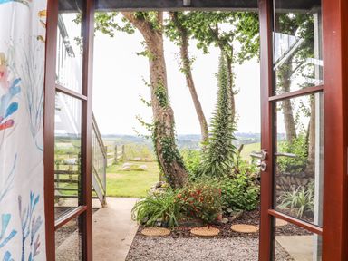 A garden view with trees plants and a grassy field seen through open glass doors at Polurrian House in Cury near Mullion
