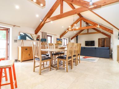 A dining area with a wooden table and chairs under wooden beams and a living area with a sofa and TV at Polurrian House in Cury near Mullion