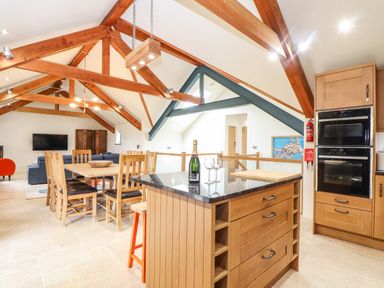 A kitchen and dining area with wooden beams wooden table and chairs a kitchen island with a bottle and glasses and built in ovens at Polurrian House Cury near Mullion