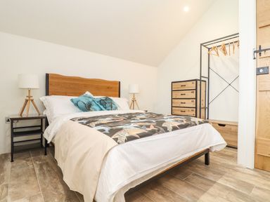 A bedroom with a wooden bed frame white bedding and two teal pillows two bedside tables with lamps a wooden dresser and an open wardrobe with hangers at Polurrian House in Cury near Mullion