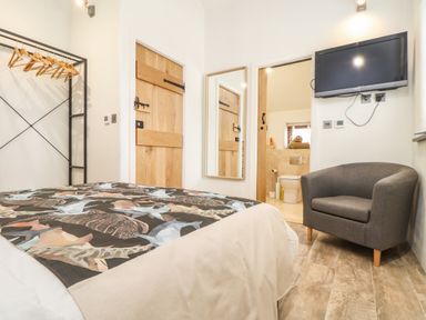 A bedroom with bed in the foreground gray armchair mounted tv wooden hanging rack mirror and door to bathroom at Polurrian House Cury near Mullion