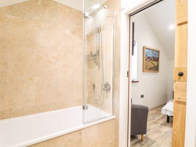 A bathroom with a bathtub and shower next to a room with a gray chair and framed painting at Polurrian House in Cury near Mullion
