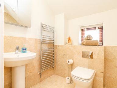A bathroom with a white pedestal sink and mirror cabinet a towel rail a toilet with a toilet paper stand and window with rolled towels at Polurrian House in Cury near Mullion