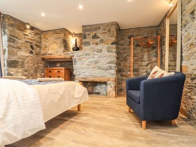 A bedroom with stone walls a wooden floor a bed a blue armchair a chest of drawers and a fireplace at Polurrian House in Cury near Mullion