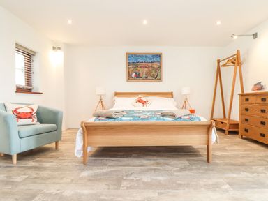 A bedroom with a wooden bed two bedside lamps a painting a blue armchair with a crab pillow a wooden dresser and an open wooden wardrobe at Polurrian House in Cury near Mullion