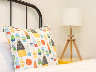 A bed with a colorful patterned cushion and a wooden tripod lamp on a yellow bedside table at Polurrian House in Cury near Mullion