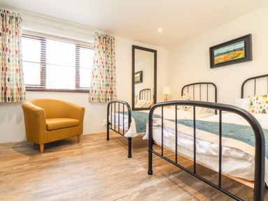 A bedroom with two single beds a yellow armchair a large mirror a lamp and a window with patterned curtains at Polurrian House in Cury near Mullion