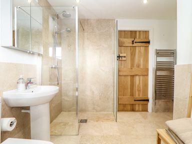 A bathroom with a glass door shower wooden door pedestal sink towel rail and folded towels on a bench at Polurrian House Cury near Mullion