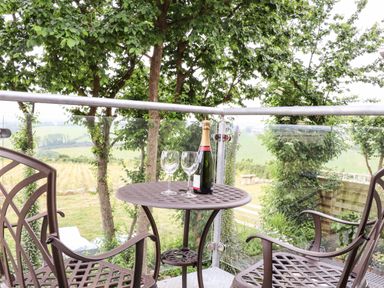 A balcony with a table holding a bottle and two glasses with two metal chairs and trees in the background at Polurrian House in Cury near Mullion