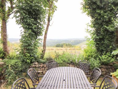 An outdoor patio with a metal table and six chairs surrounded by plants and trees overlooking a rural landscape at Polurrian House in Cury near Mullion
