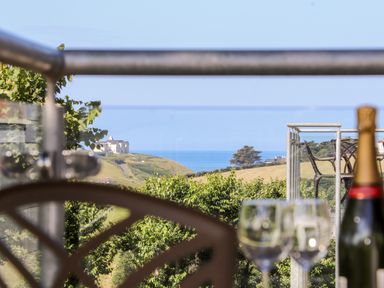 A view of the countryside with green fields and trees and a large house near the coast seen from behind a balcony railing with a table holding a bottle and wine glasses at Polurrian House in Cury near Mullion