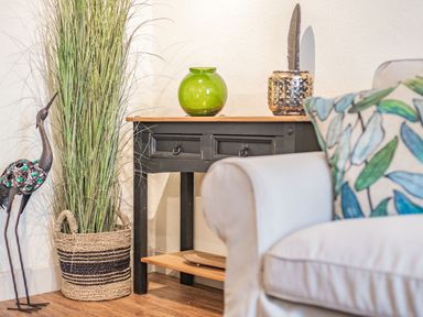 A corner with a black table holding a green vase and a decorative item beside a tall grass plant in a basket and a white sofa with a patterned cushion at Smugglers Cottage in Christchurch