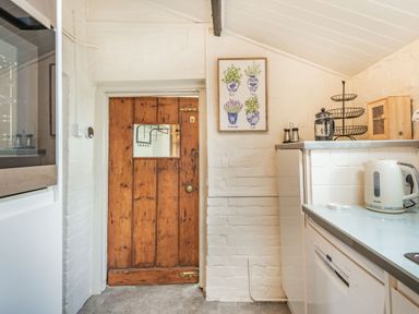 A kitchen with a wooden door framed artwork on wall electric kettle and dishwasher at Smugglers Cottage in Christchurch