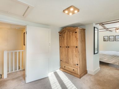 A wooden wardrobe in a carpeted room with a bed visible through an open doorway at Smugglers Cottage in Christchurch
