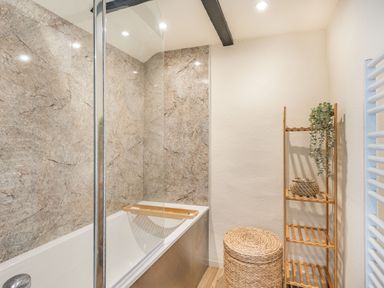 A bathroom with a bathtub and a glass shower screen with a wooden shelf a wicker basket and a wooden shelving unit with a plant at Smugglers Cottage Christchurch