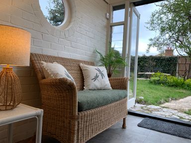 A wicker sofa with two pillows and a side table with a lamp next to an open door leading to a garden at Smugglers Cottage in Christchurch