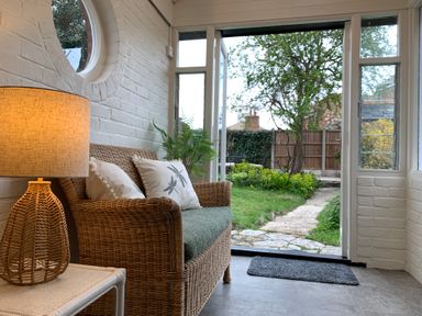 A seating area with a wicker armchair and lamp next to an open door leading to a garden at Smugglers Cottage in Christchurch