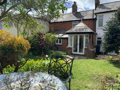 A garden with a glass table and metal chair in front of a brick house with a glass door at Smugglers Cottage in Christchurch
