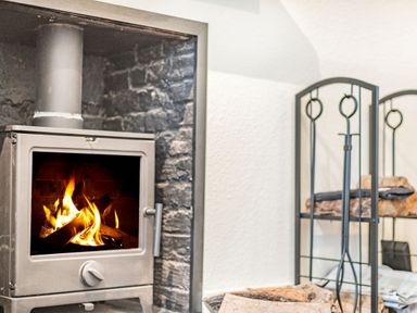 A wood burning stove with fire inside and a black metal firewood rack with firewood at Smugglers Cottage in Christchurch