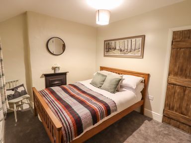 A bedroom with a wooden bed with pillows and a striped blanket beside a chair and a wall mirror at Bwthyn Y Bugail in Manod near Blaenau Ffestiniog