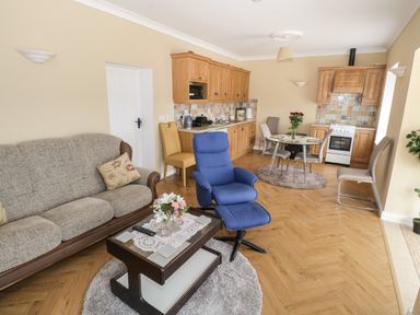 A living room with a gray sofa a blue armchair a coffee table with flowers a dining table with chairs and a kitchen area with wooden cabinets at The Water's Edge in Boyle County Roscommon