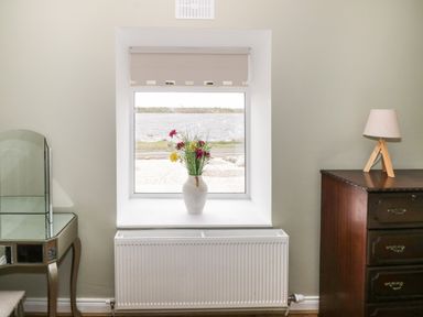 A window with a vase of flowers on the sill between a glass topped table and a wooden dresser with a lamp at The Water's Edge in Boyle County Roscommon