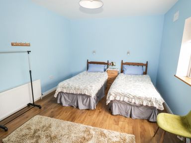 A bedroom with two single beds a wooden nightstand with flowers a rug a green chair and a clothes rack at The Water's Edge in Boyle County Roscommon