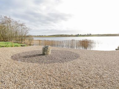 A pebble beach area with a large stone and a body of water with reeds in the background at The Waters Edge in Boyle County Roscommon