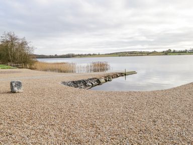 A pebble covered lakeshore with a small wooden dock and reeds in the water at The Water's Edge in Boyle County Roscommon