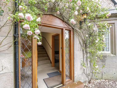 An open wooden front door with glass panes showing a staircase inside and climbing roses around the doorway at The Lodge in Greystoke