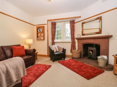 A living room with a brown leather sofa a black armchair a fireplace with a wood stove red rugs and curtains at The Lodge in Greystoke