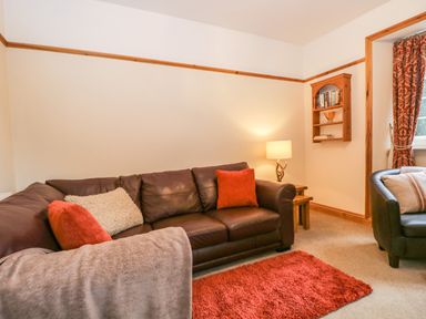 A living room with a brown leather sofa with cushions a side table with a lamp a wall shelf with books and a red rug on the floor at The Lodge in Greystoke