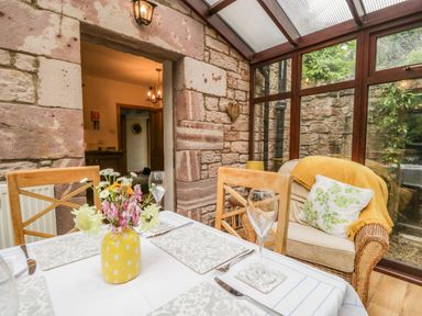 A dining table with place settings and a yellow vase of flowers next to a wicker armchair with cushions in a room with stone walls and glass windows at The Lodge in Greystoke