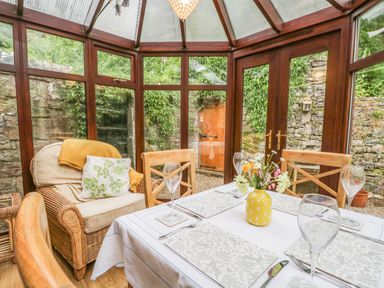 A dining area with a table set for four and a wicker armchair in a glass conservatory at The Lodge in Greystoke