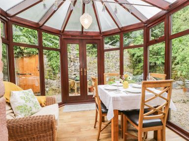 A dining area with wooden chairs and a table set with plates and glasses in a glass conservatory at The Lodge in Greystoke