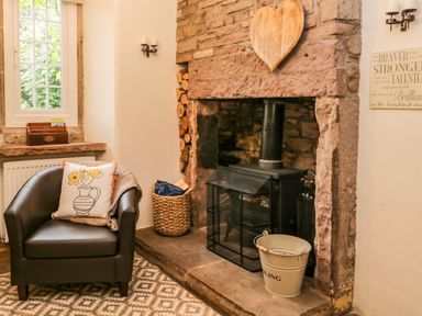 A room with a fireplace an armchair with a pillow and a blanket a basket with firewood and a window at The Lodge in Greystoke