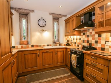 A kitchen with wooden cabinets a black stove a double sink two narrow windows and a clock on the wall at The Lodge in Greystoke