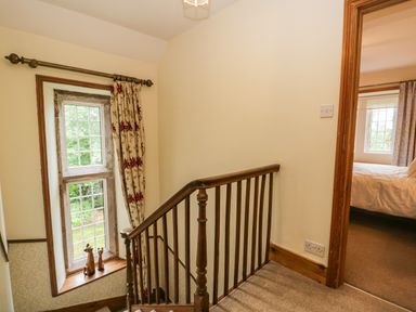 A staircase with wooden railing next to a window with curtains and a view of a bedroom at The Lodge in Greystoke