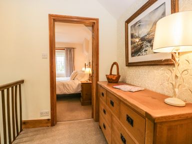 A hallway with a wooden dresser a lamp and a painting leading to a bedroom with a bed and bedside table at The Lodge in Greystoke