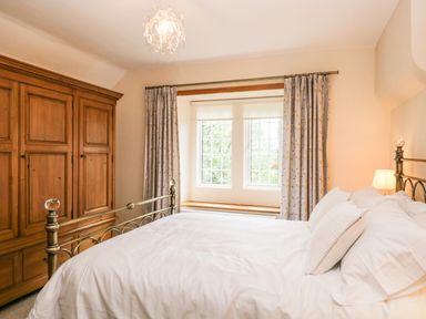 A bedroom with a white bed next to a window with floral curtains and a large wooden wardrobe at The Lodge in Greystoke