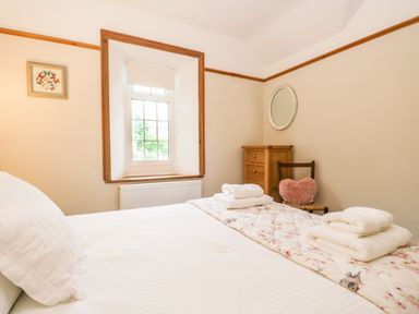 A bedroom with a bed a window wooden dresser a chair with a heart shaped cushion and folded towels at The Lodge in Greystoke