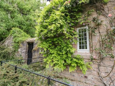 A stone building with a white window and green climbing plants next to a stone wall with a black door at The Lodge in Greystoke