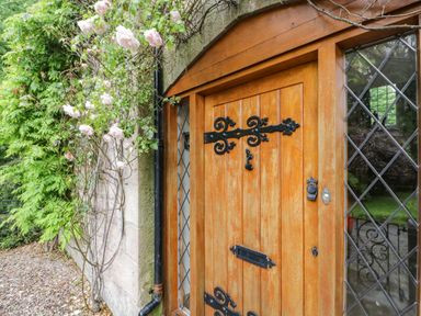 A wooden door with black metal hinges and a knocker next to glass windows with diamond panes and climbing roses at The Lodge in Greystoke