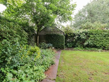 A garden with a grass lawn a paved path shrubs flowers and a small building at The Lodge in Greystoke