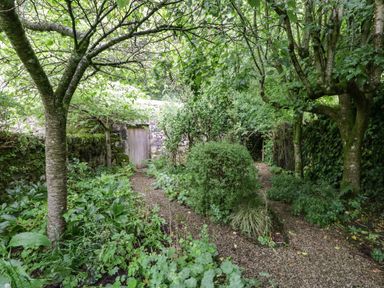 A garden with trees shrubs and a gravel pathway leading to a wooden door in a stone wall at The Lodge in Greystoke