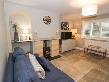A living room with a blue sofa a fireplace a television on a cabinet and a bench under a window at Dazie Cottage in Broadway